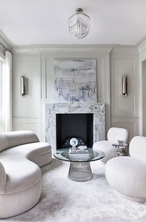 a white living room with a white marble fireplace, a glass coffee table, curved furniture, a crystal chandelier