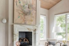 an exquisite fireplace clad with white marble, with an oversized artwork, a white chair, a floor lamp and some textiles