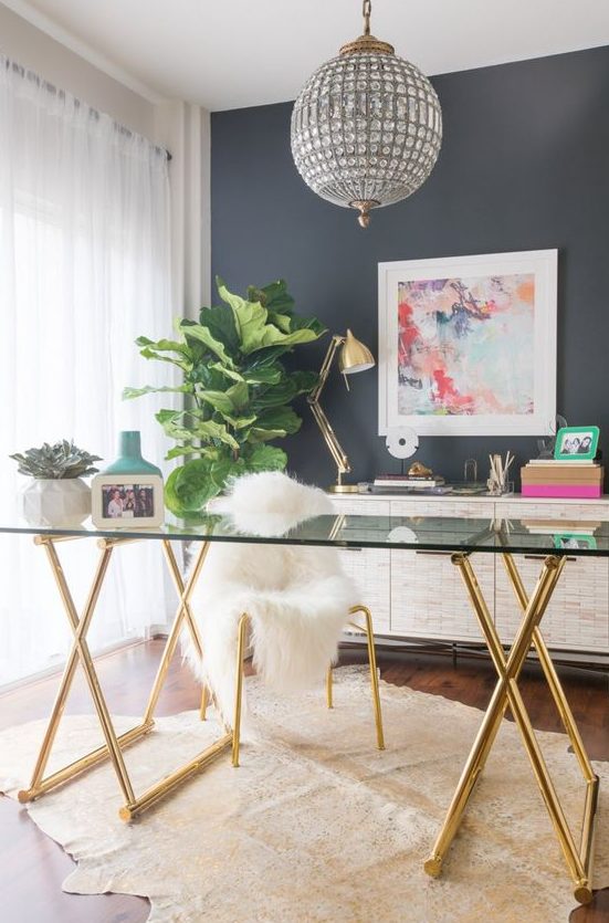 a glam feminine home office with a grey statement wall, a glass desk, a crystal chandelier and touches of gold