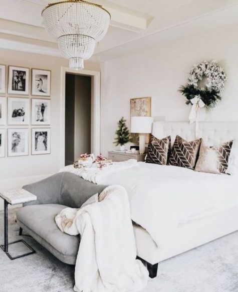 an elegant neutral bedroom with a creamy upholstered bed, a grey loveseat, a refined gallery wall, a beaded chandelier and a wreath