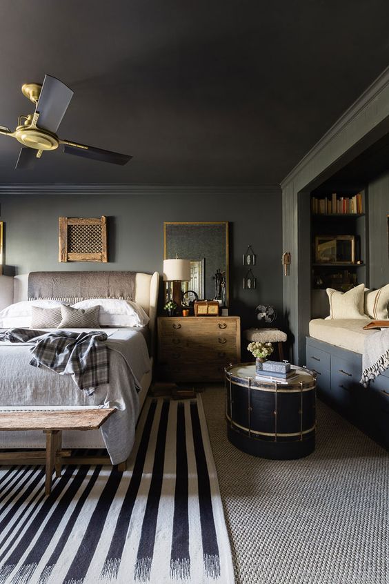 A slate grey bedroom with a cozy reading nook with built in shelves, a creamy bed with neutral bedding, stained nightstands, layered rugs