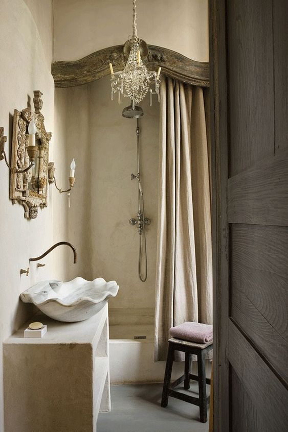 a vintage neutral bathroom with limewashed walls and a shower space, a vintage crystal chandelier, a mirror in an ornated frame and a shell sink