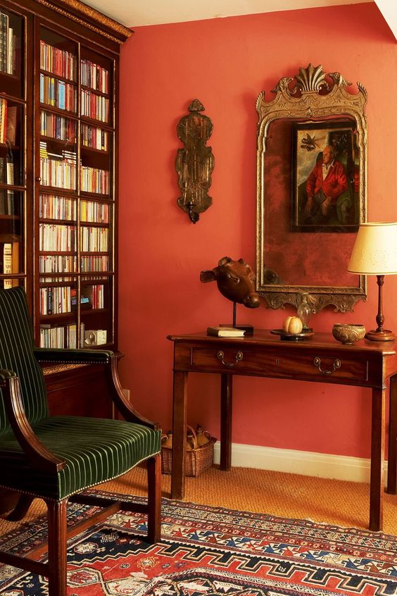 A vintage reading space with a coquelicot wall, heavy dark stained furniture, a bookcase, a mirror and vintage artwork