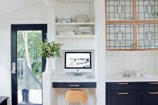 05 a farmhouse kitchen with navy cabinets, a small nook with a built-in desk, built-in shelves, a light-stained stool and potted greenery