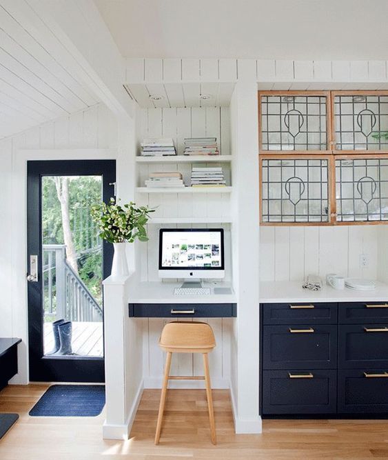 A farmhouse kitchen with navy cabinets, a small nook with a built in desk, built in shelves, a light stained stool and potted greenery