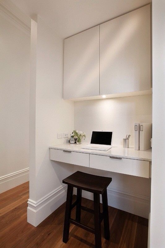 A sleek white working space with a large sleek cabient for storage, a built in desk, a dark stool and some built in lights
