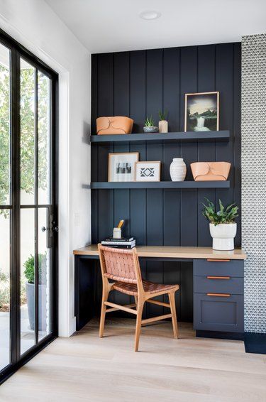 A small navy nook with shiplap, a built in desk, open shelves, a woven chair and potted plants and leather baskets