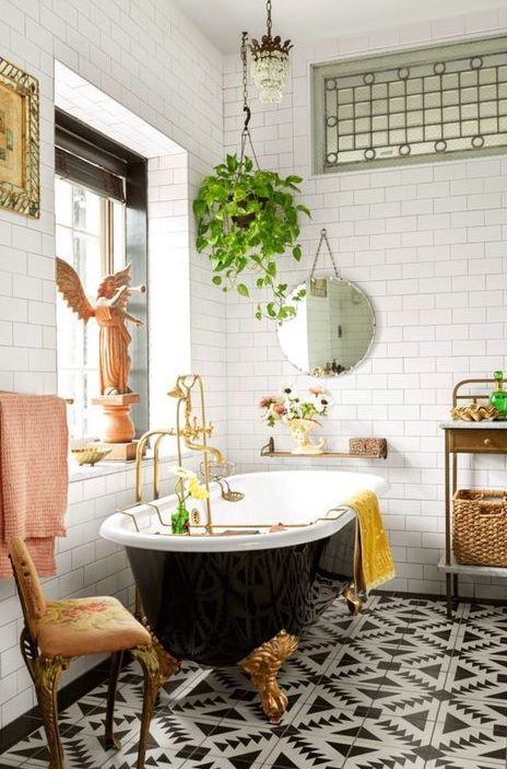 A black and white bathroom with a chic black clawfoot bathtub and bright touches   yellow and pink towels, a vintage chair, statues and potted plants