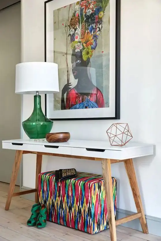 A chic white mid century modern console table with a super colorful artwork, a bright upholstered ottoman and a green lamp for summer