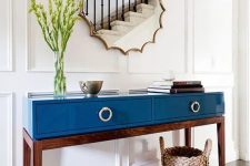 a mid-century modern table in blue lacquer finish with drawers, greenery in a clear vase, a bowl, a stack of books and a mirror of a very catchy shape