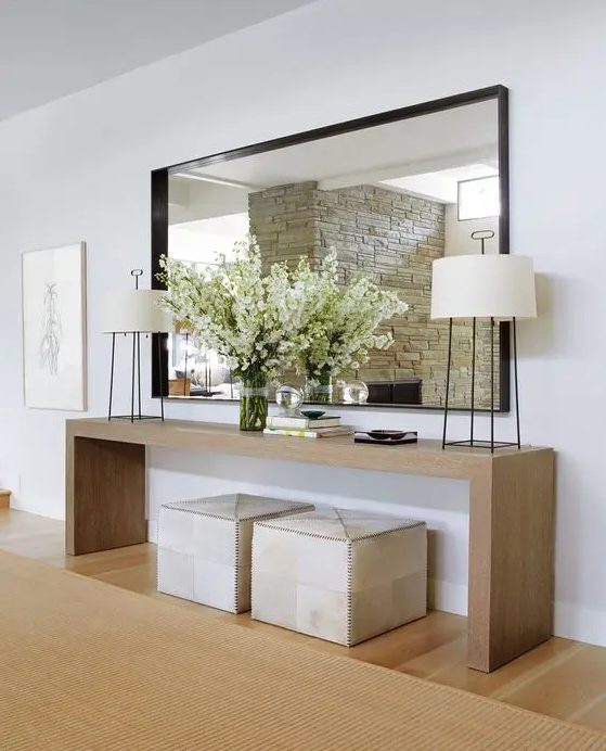 a modern sleek light-stained console table, neutral poufs, matching lamps, an oversized mirror and blooming branches in the vase