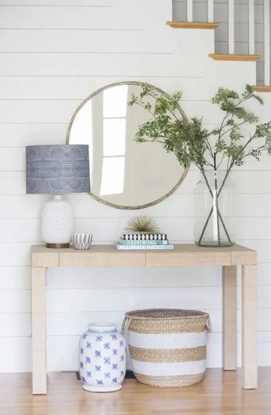 A stylish farmhouse light stained console table, a round mirror, greenery branches in a large jar, a lamp with a blue printed lampshade and a basket under the table