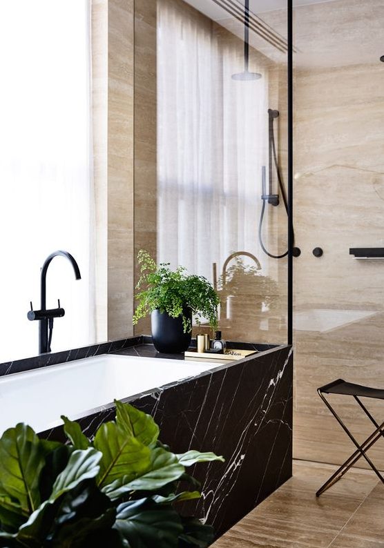 a tan stone bathroom with a shower space and a bathtub clad wiht black marble and black fixtures is a gorgeous space