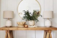 a wooden trestle leg console with a potted plant, some baskets under the console and a couple of elegant lamps plus a round mirror