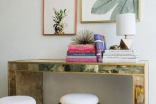 an encrusted console table, colorful books, tropical artworks over the table and hammered metal ottomans
