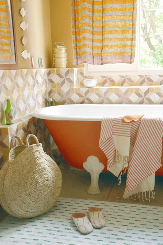 a bright bathroom with geometric tiles, an orange bathtub, a printed rug, printed textiles and natural light coming through the window