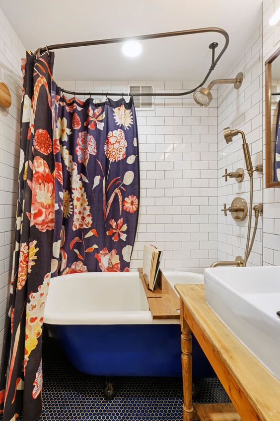a small bathroom with white subway tiles, a navy penny tile floor, a bold blue tub, a wooden vanity and a florla curtain