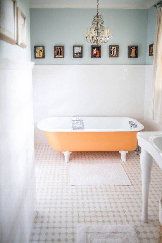 A light filled bathroom with light blue walls, an orange clawfoot bathtub, a crystal chandelier and a vintage gallery wall