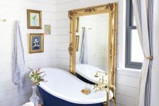 24 a refined bathroom with white shiplap, a navy clawfoot bathtub, a floor mirror in a gold frame, a crystal chandelier and artworks