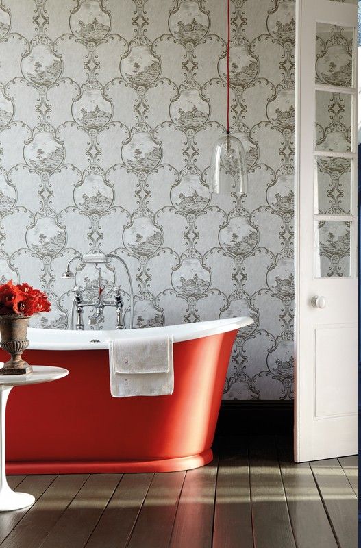A refined bathroom with grey printed wallpaper, a stained floor, a red free standing bathtub, a glass pendant lamp over the tub