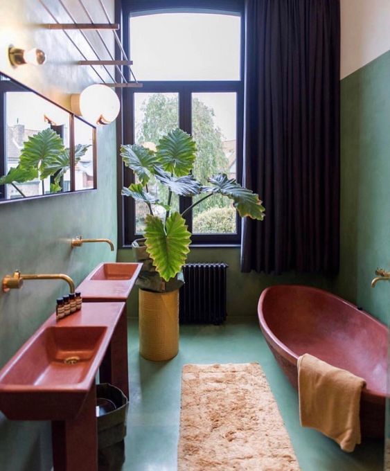 A green maximalist bathroom with red free standing sinks, a red oval bathtub, a neutral rug, potted plants and a mirror, black curtains