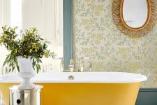 33 an elegant vintage bathroom with floral wallpaper, a mirror in a woven frame, a mustard free-standing bathtub, a shiny side table and greenery