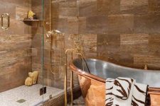 a beautiful bathroom with tan and greige tiles, a cool shower space enclosed in glass, a shiny copper bathtub with a stainless steel inner is amazing