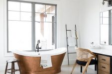 a beautiful light-filled bathroom in neutrals, with a wooden vanity and a stained and black chair, a cool copper bathtub and a ladder