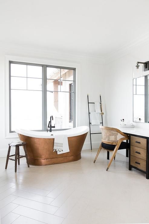 A beautiful light filled bathroom in neutrals, with a wooden vanity and a stained and black chair, a cool copper bathtub and a ladder