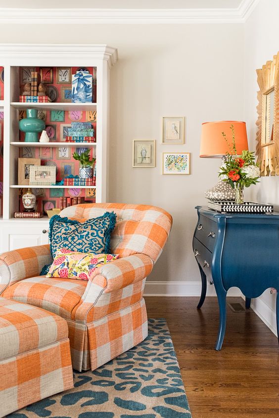 a bright living room with a bookcase with bright lining, a vintage blue credenza, a bright checked chair with a matching footrest and a mirror with a pretty frame