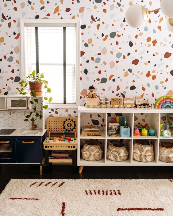 A bright mid century modern nursery with a terrazzo accent wall, a white shelving unit, a navy play kitchen, some greenery and bright toys