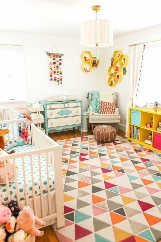 A bright mid century modern nursery with a white crib, a colorful geometric rug, a turquoise dresser, a yellow shelving unit and yellow hexagon shelves