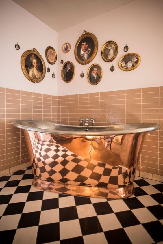 a checked black and white floor, rust tiles on the walls, a polished copper bathtub and a vintage gallery wall of oval and round artworks