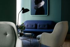 a chic and stylish living room with green walls, a navy loveseat, grey chairs, a glass coffee table and a bold and contrasting artwork