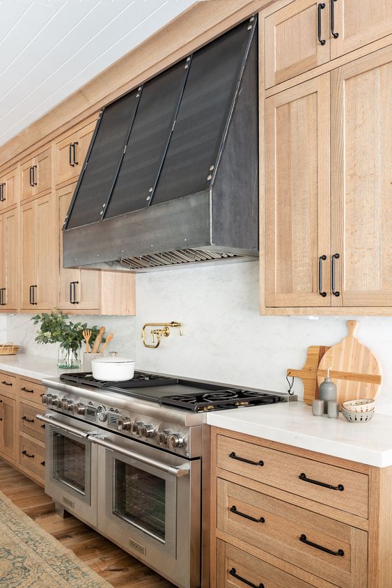 a chic light wood kitchen with shaker style cabinets, white stone countertops and a white stone backsplash, a metal hood and black handles