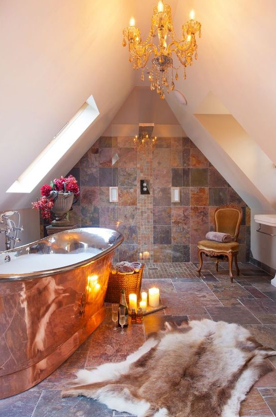 a glam attic bathroom clad with mismatching earthy tiles, with a copper bathtub and a crystal chandelier is amazing