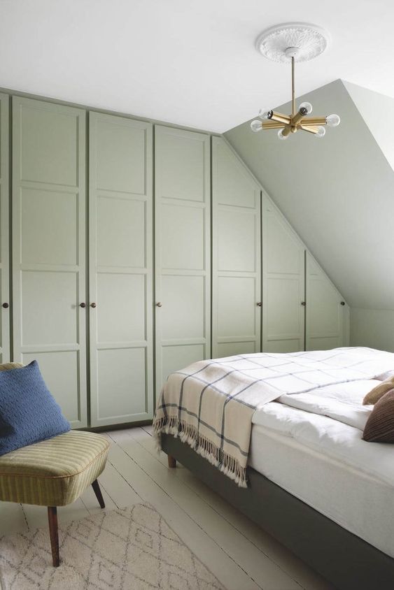 A green bedroom with built in attic and usual wardrobes, a grey bed with neutral bedding, a green chair and a rug plus an elegant chandelier