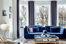 a light-filled living room wiht a bay window, a bold blue sofa, a glass coffee table and a mirror side table, a cool chair and a mid-century modern printed rug