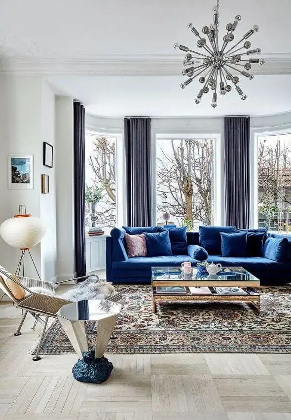 A light filled living room wiht a bay window, a bold blue sofa, a glass coffee table and a mirror side table, a cool chair and a mid century modern printed rug