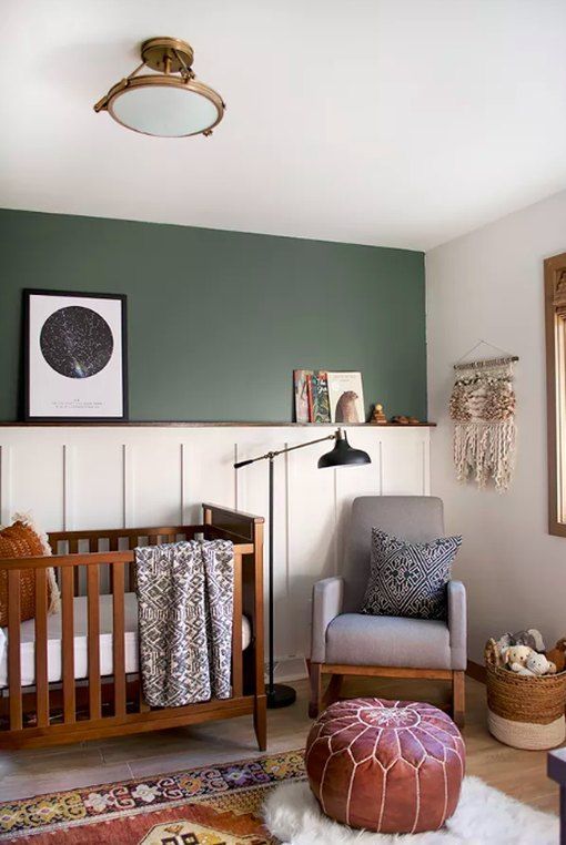 A mid century modern meets boho nursery with a green and paneled accent wall, a stained crib, a grey chair, a leather Moroccan pouf and a black floor lamp