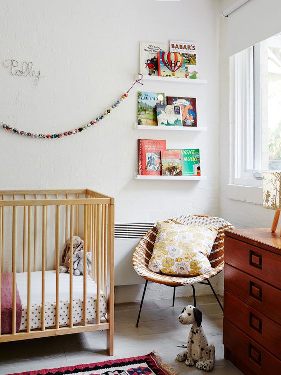 A mid century modern nursery with open shelves for books, a stained crib, a stained dresser, a wicker round chair, printed textiles and lovely toys