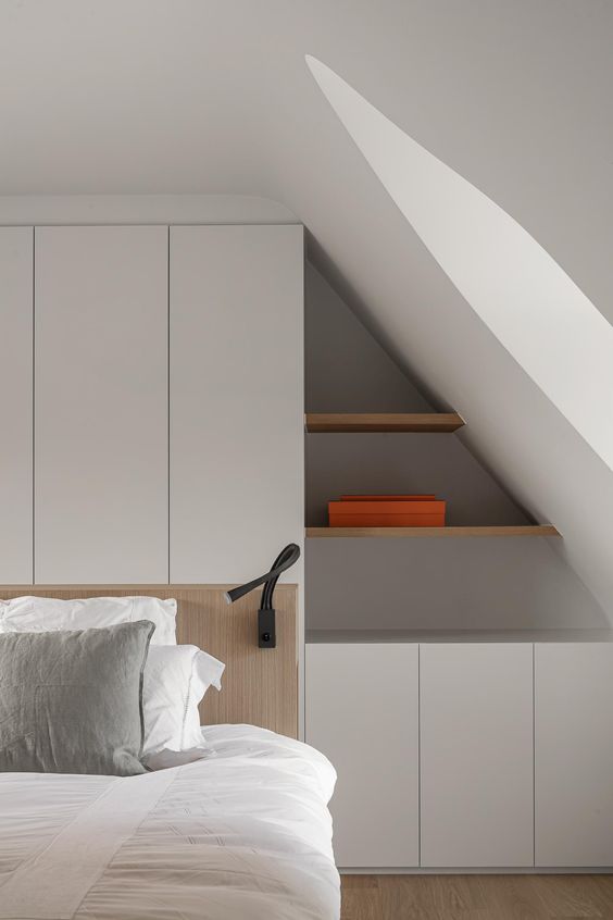 A minimalist attic bedroom with a white storage unit built into the wall, with built in shelves and cabinets is a smart solution