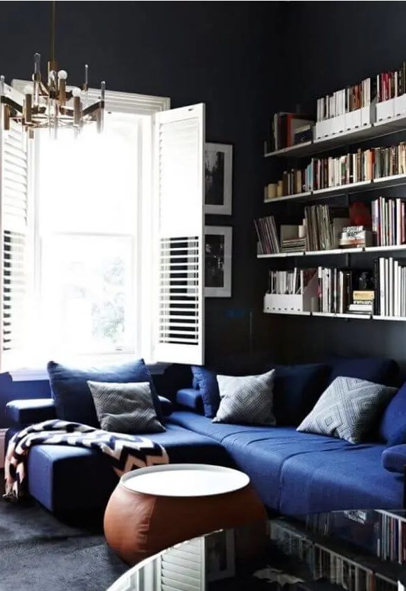 A moody living room with black walls, a navy sectional, printed pillows, open shelves, a leather coffee table, a stylsih mid century modern chandelier