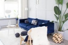 a pretty light-filled lviing room done in white and creamy, with a navy sofa, a couple of side tables, a wooden chair, a potted tree and some wood