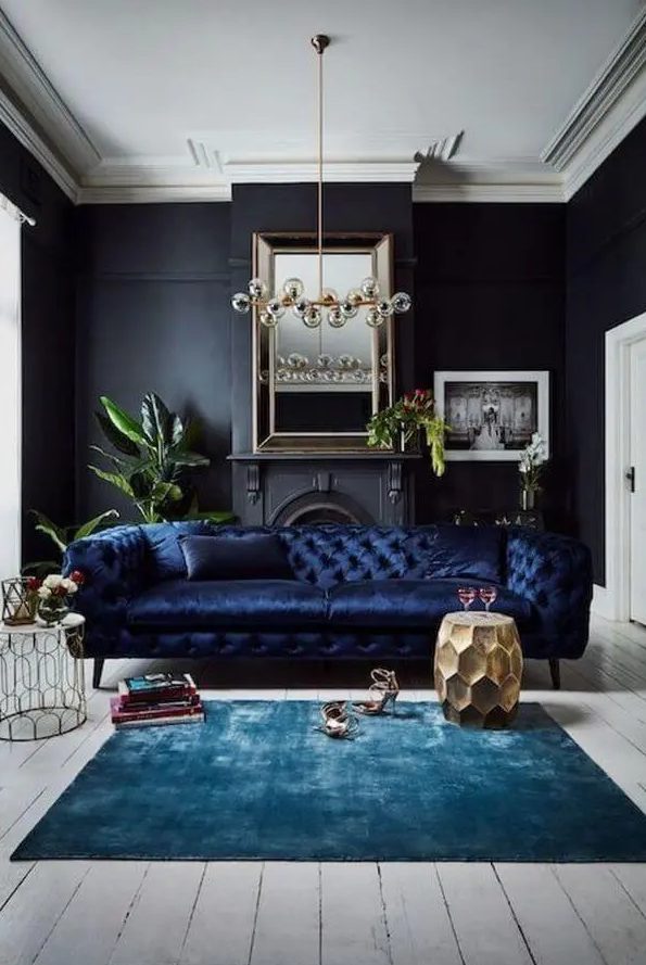 A refined living room with black walls, a non working fireplace, a navy sofa, a bulb chandelier, a turquoise rug and a gold faceted side table and some books and plants