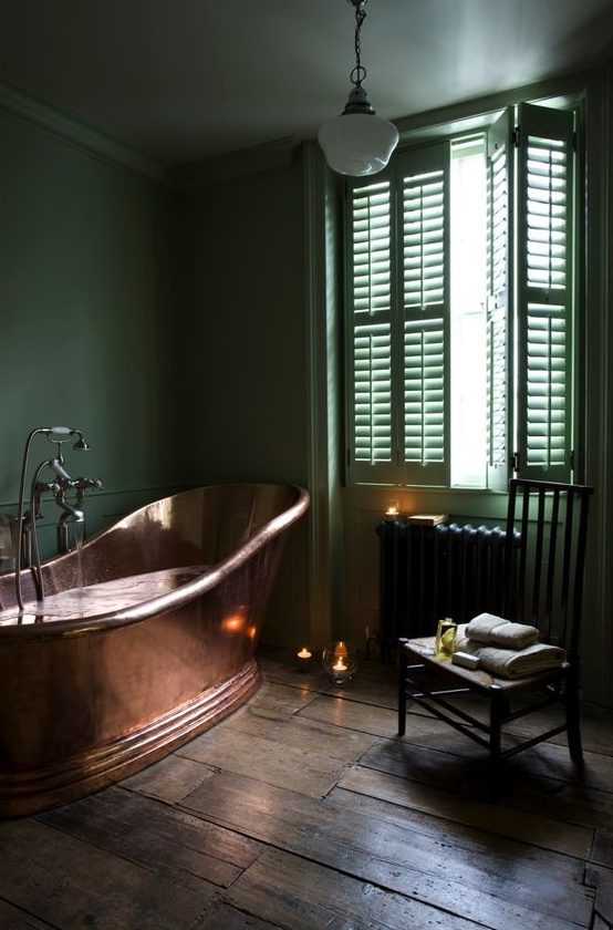 a refined moody bathroom in green, with a black radiator, a copper bathtub and candles around is very chic