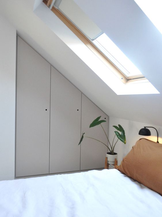 a small attic bedroom with a built-in storage unit with grey doors, a bed, a black lamp and a potted plant plus some skylights