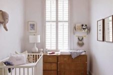 a small modern nursery in neutrals, with a white crib, a stained dresser as a changing table, a basket with toys, a table lamp and artworks