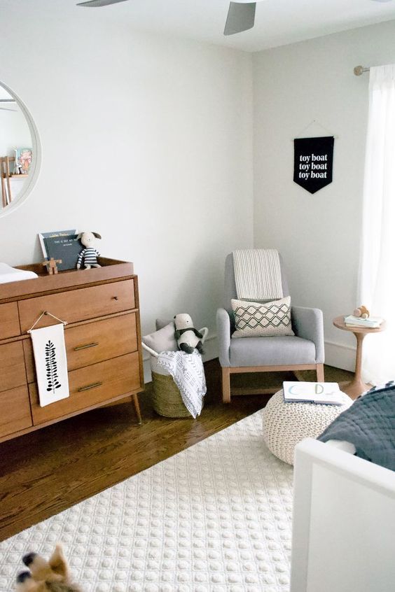 A welcoming mid century modern nursery in neutrals, with a grey rocker, a stained dresser, a white crib, a white pouf, baskets with toys and printed textiles