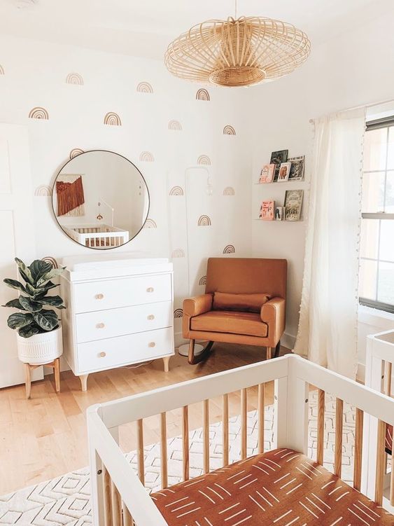 an earthy tone mid-century modern nursery with pretty wallpaper, a white dresser, an amber leather chair, a white crib, a wooden pendant lamp and books